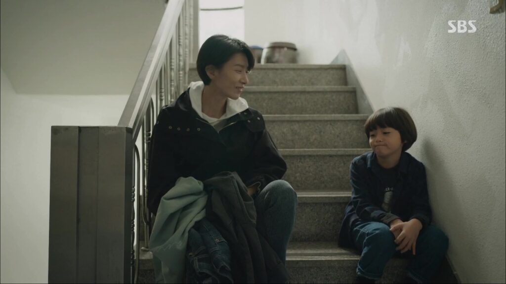 Cha-young and a young boy sit on the stairs of an apartment building. She is looking at him with affection