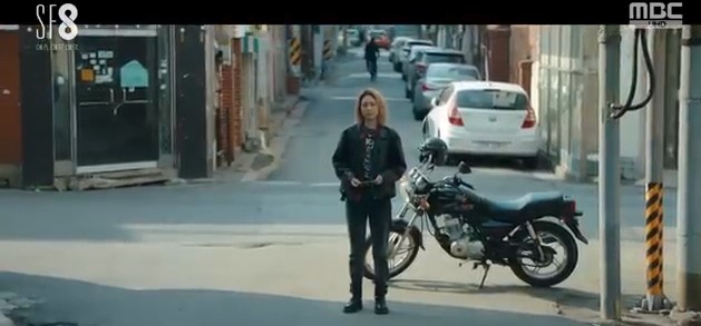 Lead character To Sun-ho, dressed in leather on a city street with her motorcycle parked behind her