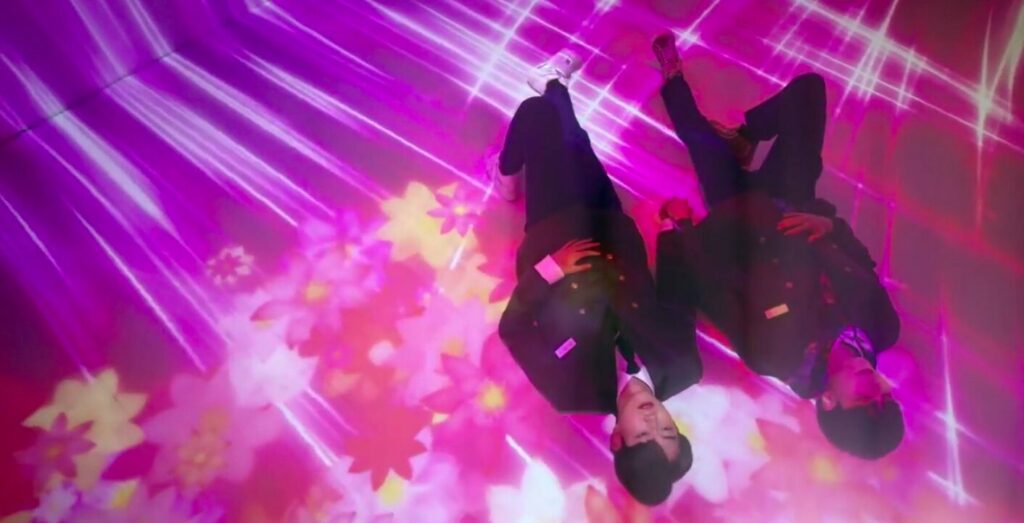 The two male leads in Colour Rush in a multimedia prsentation about colour. They're lying on the ground completely surrounded in bright powerful colours like purplle