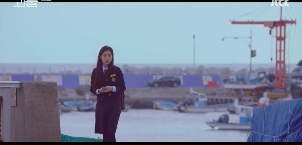 A teenaged Jae-yi in a school uniform stands on the docks in Busan looking lost as she searches for her mother