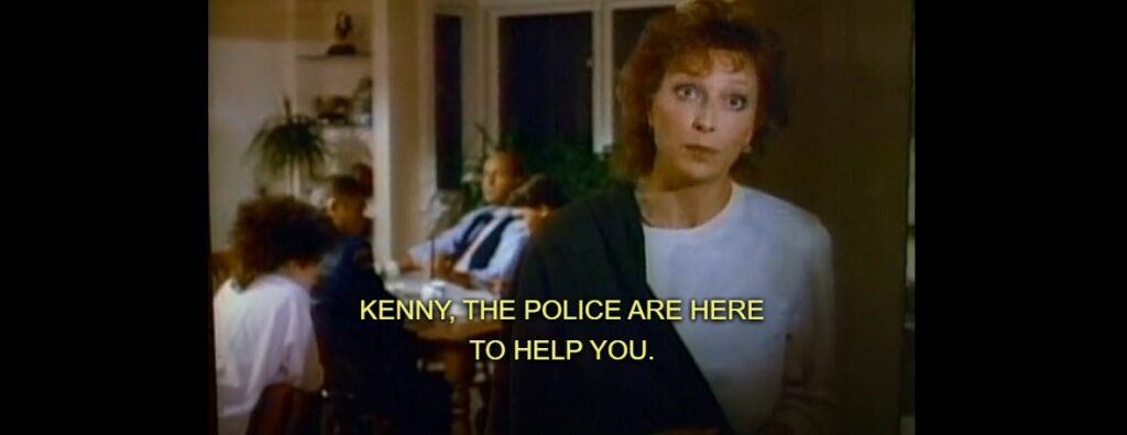 The mother from the beginning scene stands at the bottom of a flight of stairs and yells up to her son who's not in frame, "Kenny, the police are here to help you"