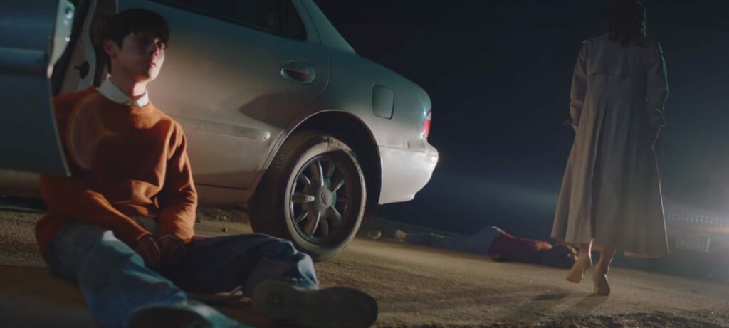 Jung-jae sits on teh ground by his car. The door is open and he's framed in the headlights of another car. His mother moves away from him towards the body of a young woman lying on the ground in the road