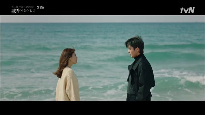 The two leads of DAYS swap meaningful looks on the beach while the endless ocean stretches out behind them