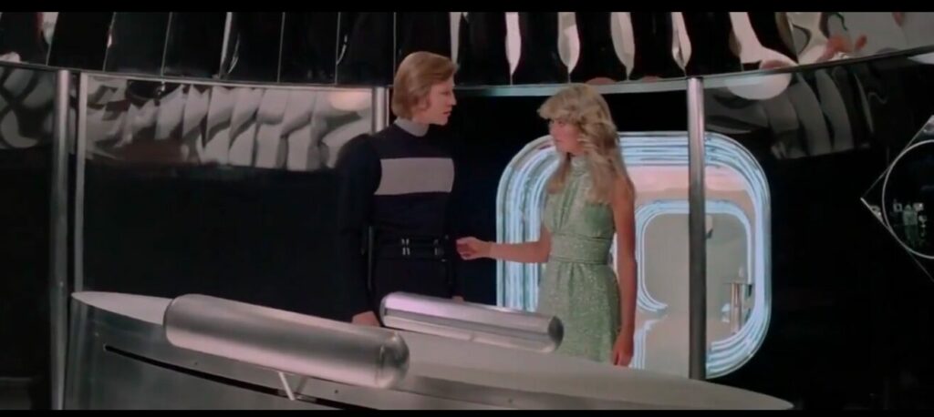 Logan standing next to a futuristic plastic surgery table with an attendant who has Farah Fawcett hair -because she's literally Farah Fawcett