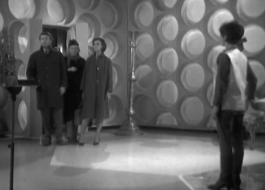 Ian and Barbara stand in a large white room with roundels on the walls. The Doctor is coming through the door behind them and Susan stands by the console