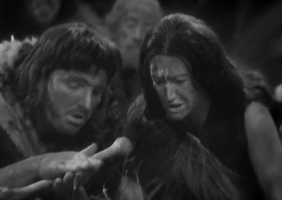 A paleolithic man tries to start fire with a bone while a woman looks on, concerned.