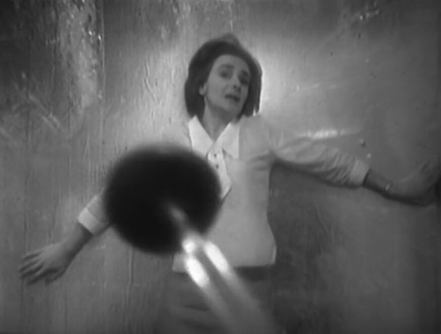 Barbara reels back in terror against a metal wall while a plunger comes at us menacingly