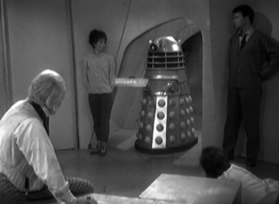 The Daleks bring a tray into the prisoners' cell. Susan and Ian stand on either side of the door. Barbara is on the floor and the Doctor sits on a bench. It's a trap for the Dalek. 