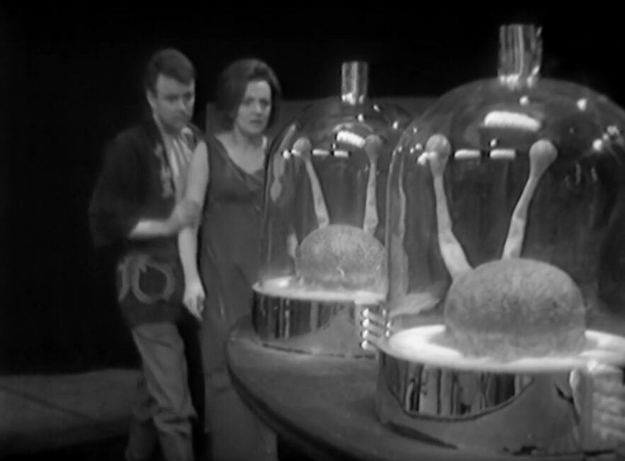 Ian and Barbara stand horrified and transfixed by two glass jars with a lump of brain matter with two eye stalks protruding from them