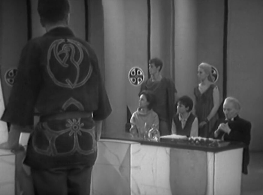 Ian stands with his back towards us at a table while nearby Barbara, Susan and the Doctor sit at a defence table looking concerned