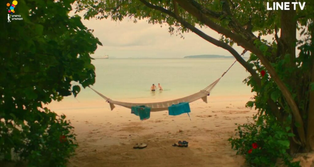 A hammock between two trees frames the shot. Two shirts hang off it and two pairs of shoes are below it. Two boys in the ocean walk towards it