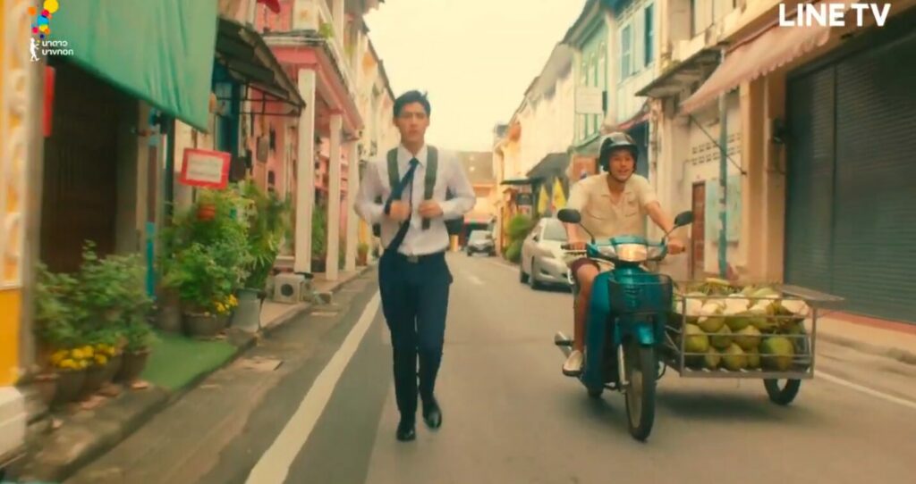 Oh-aew runs down a street in his school uniform, Teh beside him on a motorcyle with a side car. The street is lined by multi-coloured colonial buildings