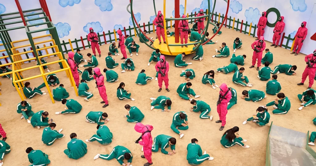 Men in pink jumpsuits holding guns guard a group of contestants in green tracksuits chipping away at honeycomb in a giant adult-sized playground.