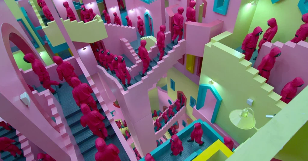 A surrealistic landscape inside the Squid Game. Bright pastel staircases in pink, blue and green criss cross the image seemingly going nowhere like Escher stairs. On the stairs are dozens of people in pink jumpsuits with black masks.