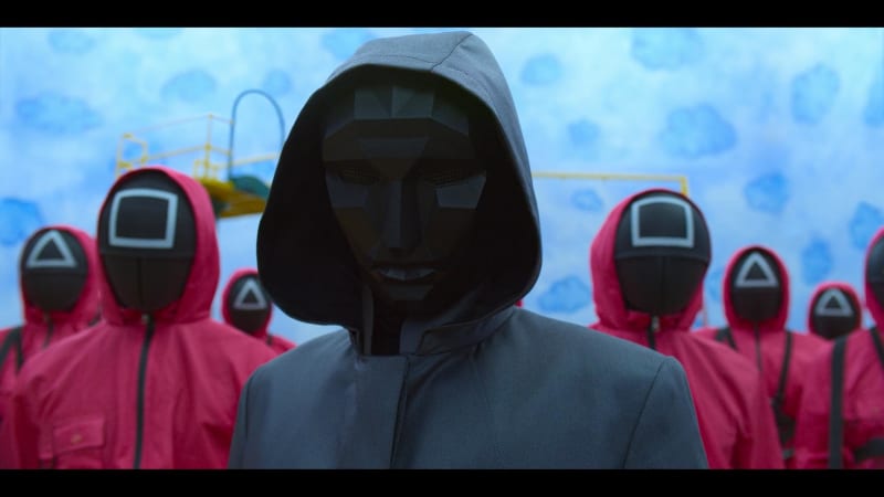 A man in a black mask and a black hooded coat stands in front of a group of men dressed in pink jumpsuits. They have black masks with geometric shapes on the front in clear white lines.