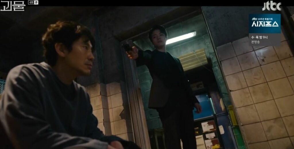 Dong-shik sits in his basement. Standing over him is Joo-won pointing a gun at his head
