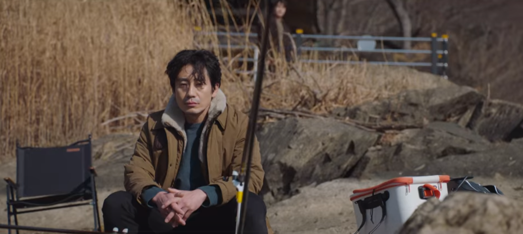 Shin Ha-kyun as the male lead in Beyond Evil sitting by the water with a fishing pole and a look of devestation on his face