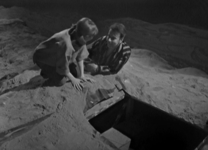 Ian and Vicki crouch on the sand behind a hatch into the ground and debate whether to climb into the dark hole on a strange planet while the sun is about to set.