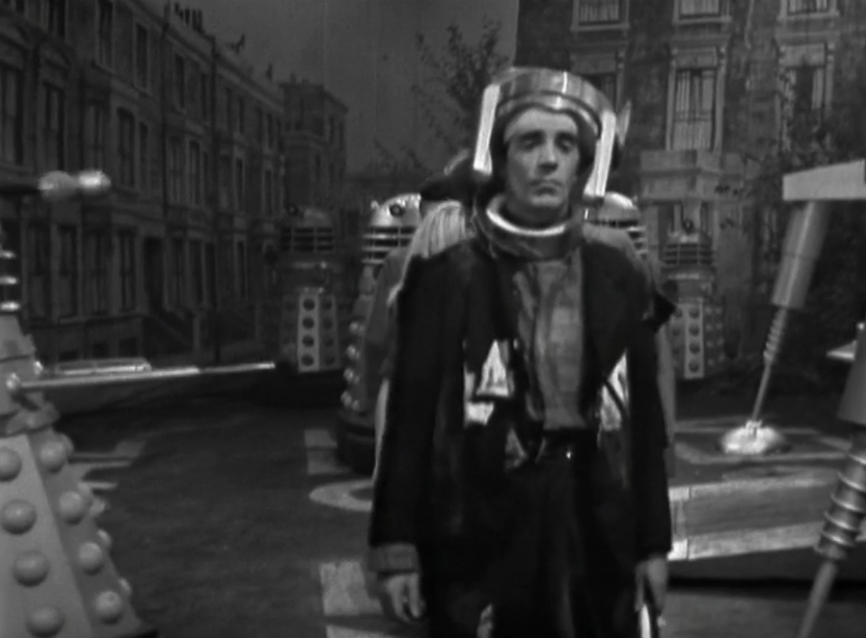 A lean lurching man with a metal helmet walks through the streets of London flanked by Daleks
