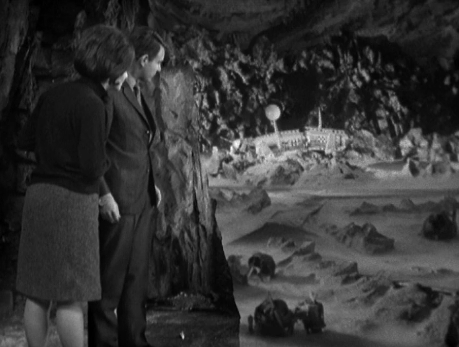 Ian and Barbara stand overlooking a ruined city on a barren planet below