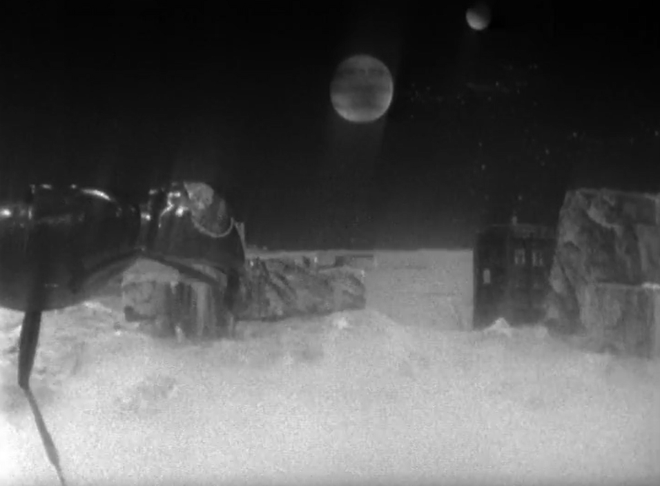 An alien landscape, barren like the moon. There is a planet in the sky and the TARDIS is tucked behind a large rock while a huge ant looks on.
