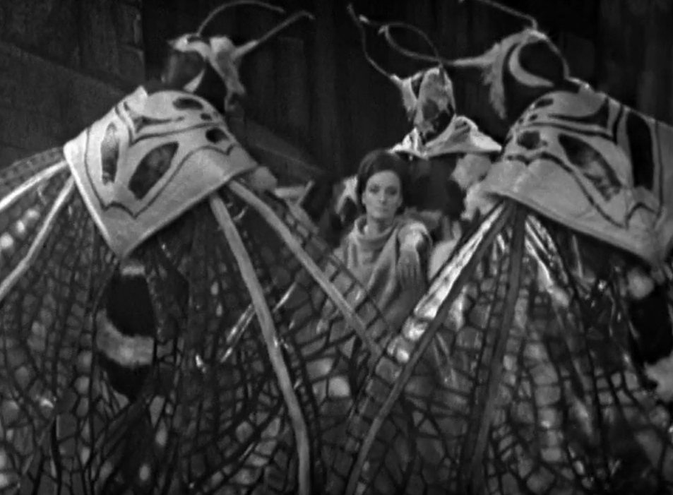 An entranced Barbara with her hand outstretched with the gold band on it is surrounded by three giant humanoid butterlies with antennas and wings.