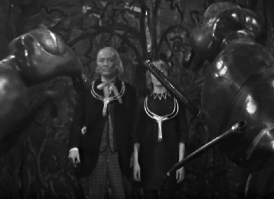 Flanked by giant ants, the Doctor and Viki wear gold collars around their necks and look hypnotised.