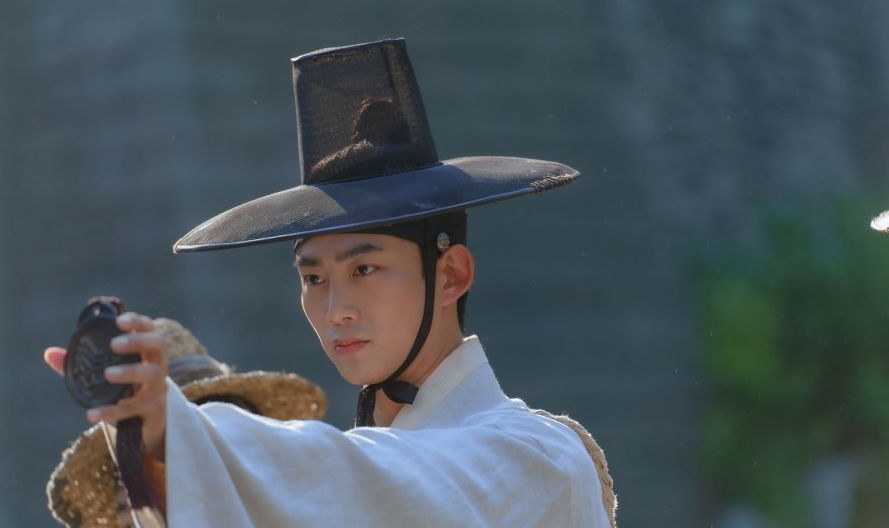 Taecyeon from 2PM holds up his Royal Ispector seal. He's wearing a gat. His ears stick out.