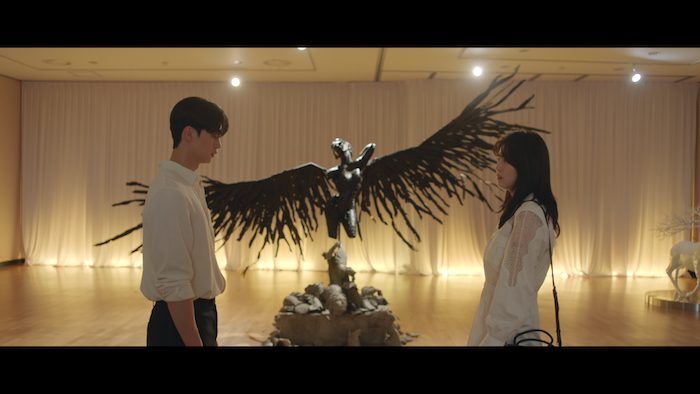 A man and a woman stand staring at each other. A statue with wings outstretched stands between them boht bridging and separating them